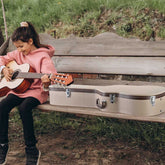 Child playing guitar next to an open guitar case on a wooden bench outdoors.