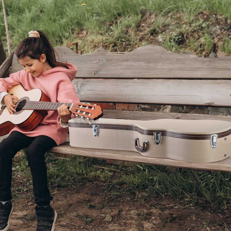 Child playing guitar next to an open guitar case on a wooden bench outdoors.