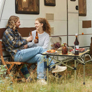 Two people sitting at a table outdoors next to a camper van, enjoying drinks and food.