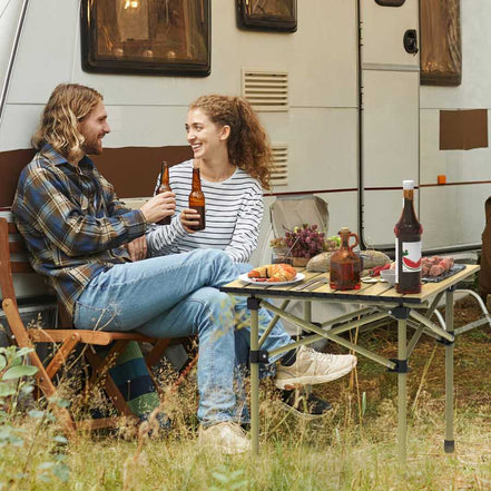 Two people sitting at a table outdoors next to a camper van, enjoying drinks and food.