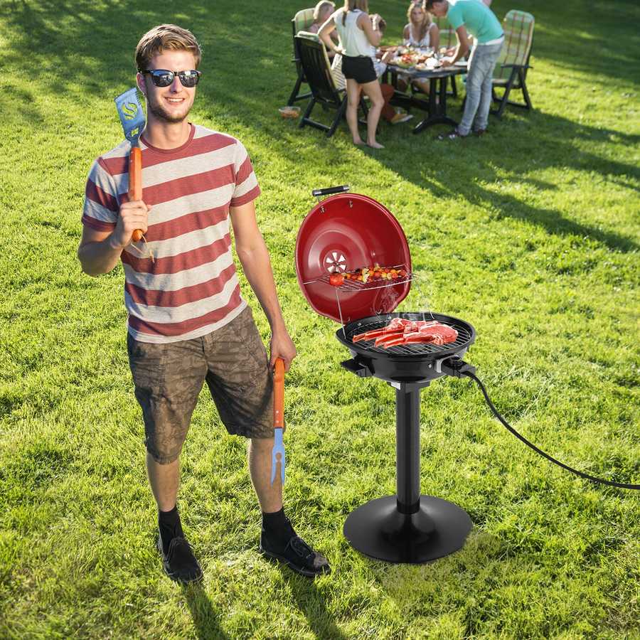 Man holding a spatula and standing next to a portable grill on grass with people in the background.