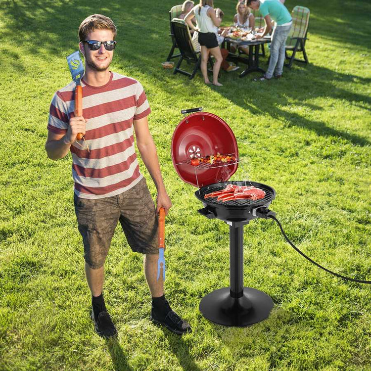 Man holding a spatula and standing next to a portable grill on grass with people in the background.