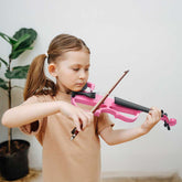 Young girl playing a pink violin indoors with a plant in the background