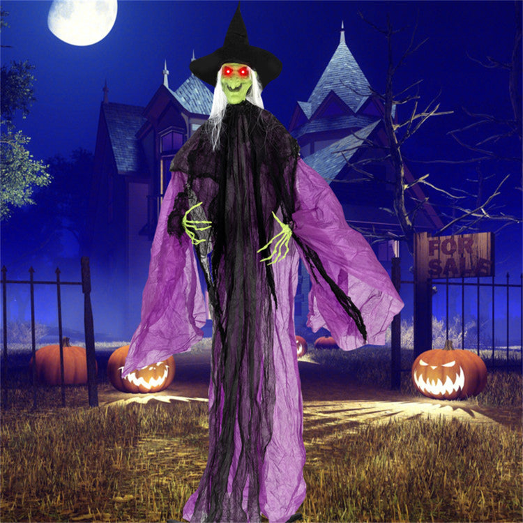 A tall inflatable Halloween witch decoration stands in a spooky yard at night. The figure wears a black hat and long black dress with flowing purple cape panels, green skin, and red glowing eyes.