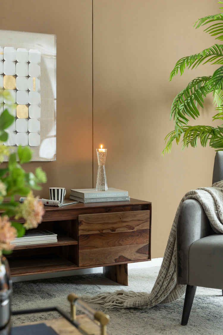 A warm, modern living room scene featuring a dark wood sideboard console with a lit candle and books, set against a beige wall with a gray armchair and a green houseplant.