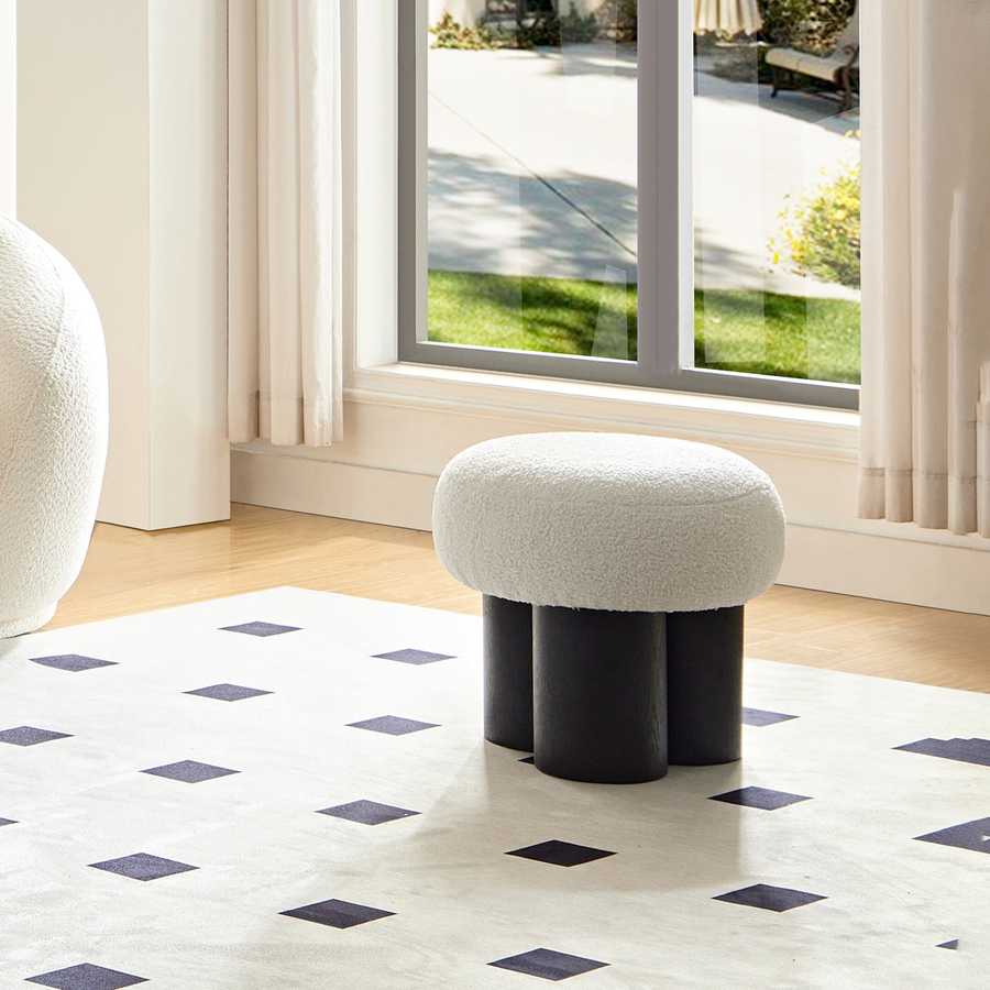 White ottoman with black legs on a patterned rug in a room with large windows.