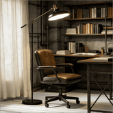 A brown leather office chair with a black metal base and five wheels, positioned in a styled home office next to a black floor lamp with a brass lampshade.