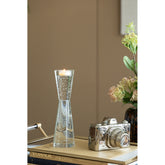 A clear glass hourglass-shaped candle holder with a lit candle inside, styled on a tabletop next to a silver camera and books in a warm indoor setting.