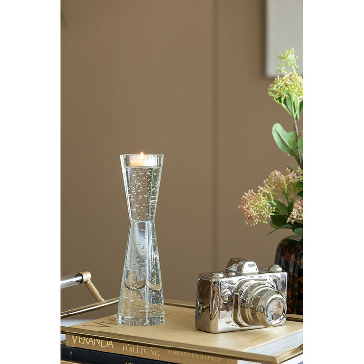 A clear glass hourglass-shaped candle holder with a lit candle inside, styled on a tabletop next to a silver camera and books in a warm indoor setting.
