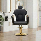 A modern black salon barber chair with gold accents and a gold-plated circular base, set in a bright, contemporary salon interior with light wood floors and white cabinets.