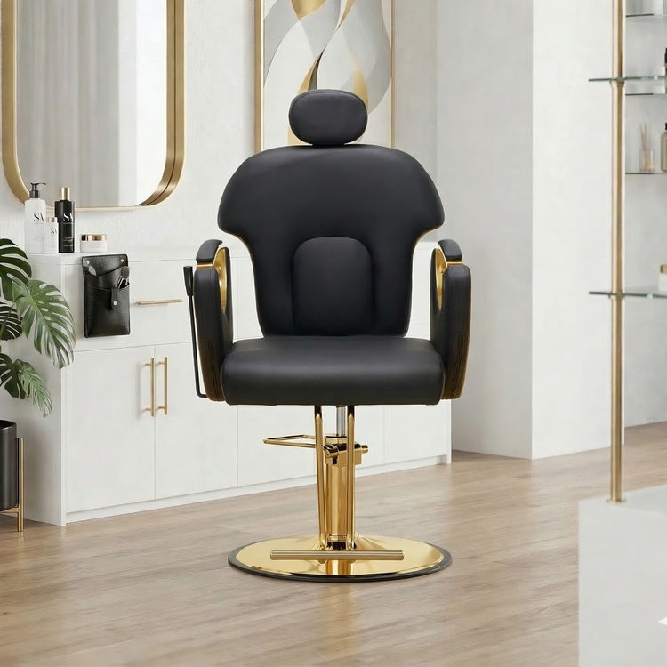 A modern black salon barber chair with gold accents and a gold-plated circular base, set in a bright, contemporary salon interior with light wood floors and white cabinets.