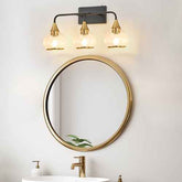 A three-light wall sconce with warm brass and black metal hardware and clear glass shades, mounted above a round gold-framed mirror in a bathroom setting.