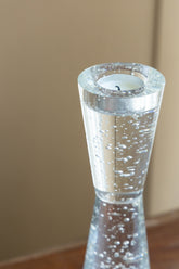 A clear glass candle holder with a conical shape filled with small bubbles, holding a white tea light candle, set against a warm beige background.