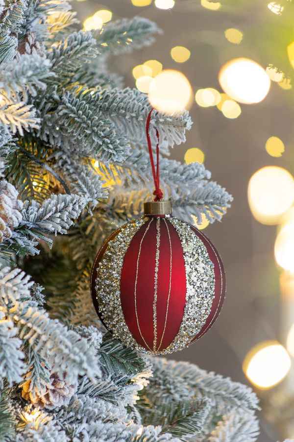 Red ornament with gold stripes hanging on a decorated Christmas tree with blurred lights in the background.