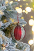 Red ornament with gold stripes hanging on a decorated Christmas tree with blurred lights in the background.
