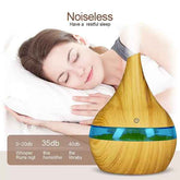 A teardrop-shaped wooden diffuser with a blue band and green liquid inside, shown in front of a sleeping woman on a white pillow. Text at the top reads &
