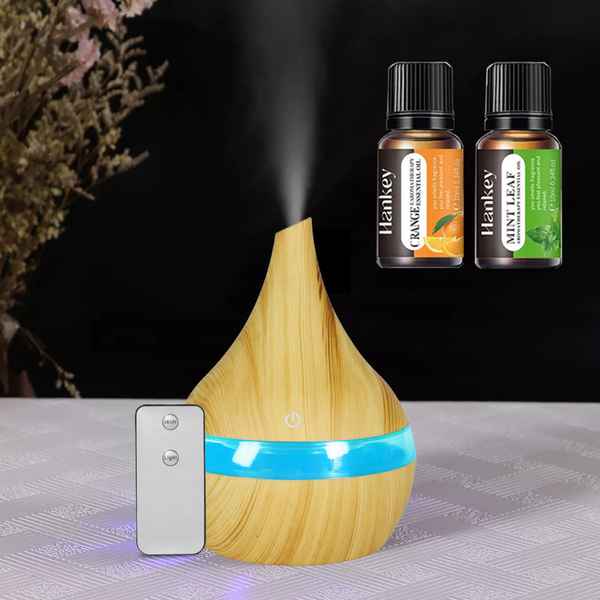 A wooden diffuser with a glowing blue band and misting nozzle, shown on a tabletop with a remote control and two small Hankey essential oil bottles against a dark background.
