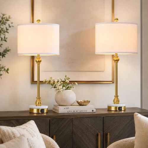 Two matching table lamps with white drum shades and polished gold bases are placed on a dark wood console in a styled interior setting with a mirror, books, flowers, and cushions visible.