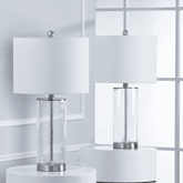 Two matching table lamps with white drum shades and clear textured glass cylindrical bases, displayed on white round pedestals in front of a white paneled wall and window.