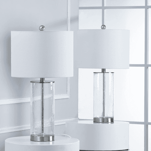 Two matching table lamps with white drum shades and clear textured glass cylindrical bases, displayed on white round pedestals in front of a white paneled wall and window.
