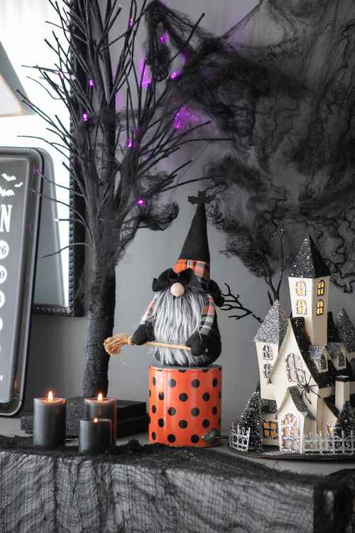 A Halloween-themed tabletop display featuring a gnome figurine sitting on an orange pot with black polka dots, next to a miniature haunted house, black candles, a spooky tree with purple lights, and cobweb decorations against a gray wall.
