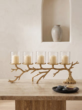 A five-light branch-shaped candle holder in a natural wood finish, styled on a stone table with five clear glass candle holders containing cream pillar candles. A small black plate with decorative stones and a book sits in the foreground, and a neutral-toned ceramic vase is displayed in the background on a recessed wall shelf.