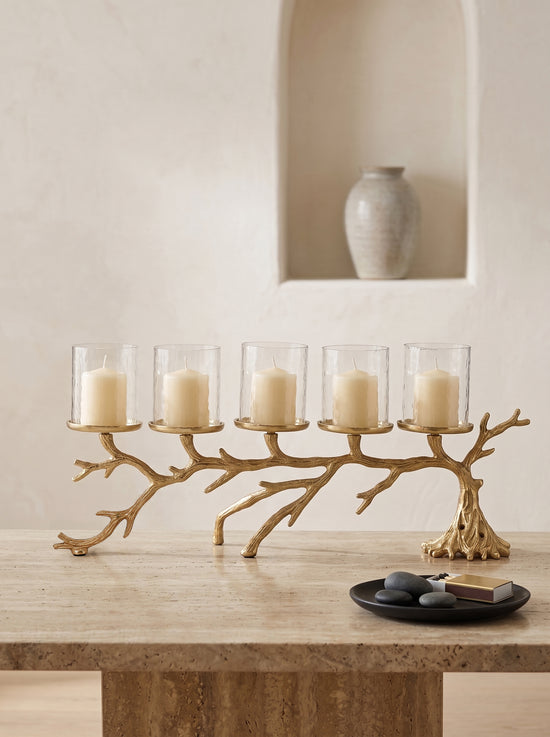 A five-light branch-shaped candle holder in a natural wood finish, styled on a stone table with five clear glass candle holders containing cream pillar candles. A small black plate with decorative stones and a book sits in the foreground, and a neutral-toned ceramic vase is displayed in the background on a recessed wall shelf.