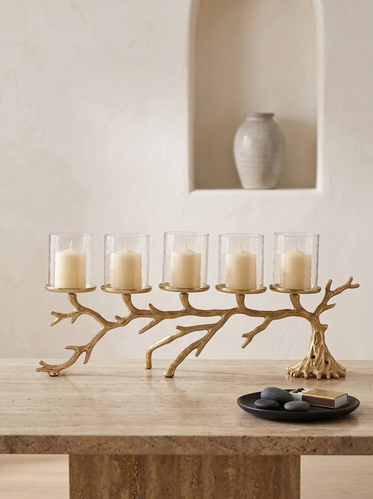 A five-light branch-shaped candle holder in a natural wood finish, styled on a stone table with five clear glass candle holders containing cream pillar candles. A small black plate with decorative stones and a book sits in the foreground, and a neutral-toned ceramic vase is displayed in the background on a recessed wall shelf.