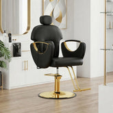 A modern salon barber chair in black faux leather with polished gold metal accents and a gold hydraulic base, set in a bright, contemporary salon interior with light wood floors and white cabinets.