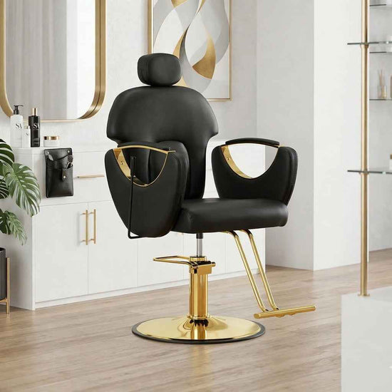 A modern salon barber chair in black faux leather with polished gold metal accents and a gold hydraulic base, set in a bright, contemporary salon interior with light wood floors and white cabinets.