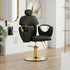 A modern salon barber chair in black faux leather with polished gold metal accents and a gold hydraulic base, set in a bright, contemporary salon interior with light wood floors and white cabinets.