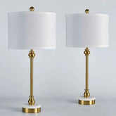 Two matching table lamps with white drum shades, polished gold metal stems and finials, and round white marble bases, shown on a light gray studio background.