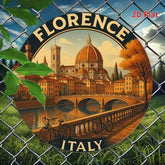 A circular printed sign or sticker featuring an illustrated Florence scene with the Duomo dome, bridge, river, bicycles, and the words “FLORENCE” and “ITALY.” The sign is shown behind a chain-link fence with grass and leaves in front.