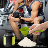 A fitness scene in a gym with a seated man lifting a dumbbell while a hand scoops white powder from a black tub with a green scoop. The tub has a black body with a bright green label, and a pile of white powder sits on the floor in the foreground.