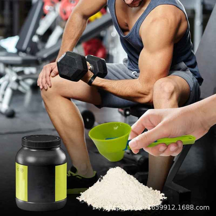 A fitness scene in a gym with a seated man lifting a dumbbell while a hand scoops white powder from a black tub with a green scoop. The tub has a black body with a bright green label, and a pile of white powder sits on the floor in the foreground.