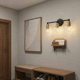 A modern wall-mounted three-light sconce with a black metal frame and warm brass/gold-toned shades, shown lit in a hallway entryway above a wooden bench and shelf.