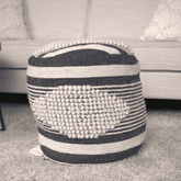 A round woven pouf ottoman with navy blue and cream stripes and a large textured cream tufted center, placed on a light gray rug in front of a light beige sofa.