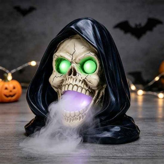Halloween decoration of a hooded grim reaper skull with glowing green eyes and a purple glowing mouth, with white smoke rising, set on a wooden surface with Halloween decorations in the background.