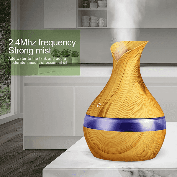 A wooden-texture essential oil diffuser and humidifier with a purple band around the middle, shown on a kitchen counter with mist coming out. Text on the left reads &