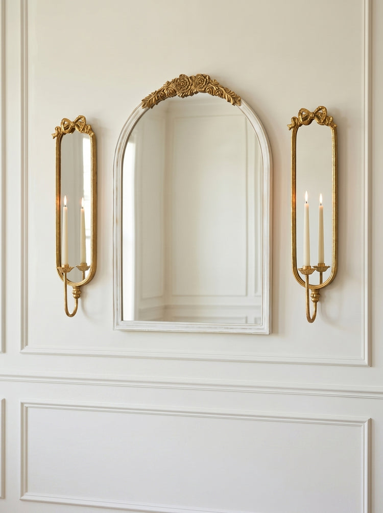 A decorative wall mirror with an ornate gold top frame, flanked by two matching gold sconces with candle-style lights, mounted on a white paneled wall.