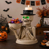 Halloween skeleton candle holder figurine holding a bowl of candy, with a lit white candle on its black hat, set on a dark wooden table in a decorated Halloween room.