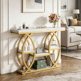 A modern console table with a white marble-look top and shelf surfaces, supported by a gold-tone metal frame with interlocking circular arches. The table is styled in a bright living room with decorative items, a floral arrangement, and a sofa in the background.