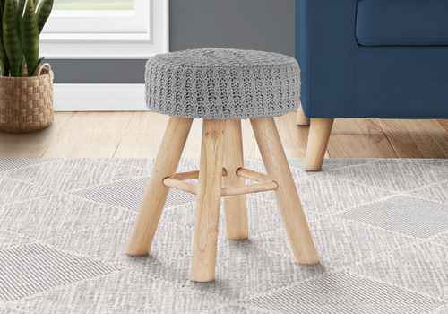 A small round stool with a gray knitted cushioned seat and natural wood legs, shown in a styled room setting on a patterned rug next to a blue armchair.