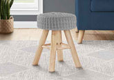 A small round stool with a gray knitted cushioned seat and natural wood legs, shown in a styled room setting on a patterned rug next to a blue armchair.