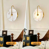 A split comparison of a modern wall sconce in a bedroom, showing the light off on the left and on the right. The sconce features a round white marble-look shade with gold trim and a curved gold metal arm.