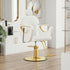 A modern salon barber chair with a white upholstered seat and gold metal armrests, backrest details, and a gold hydraulic base, shown in a bright, contemporary salon interior.