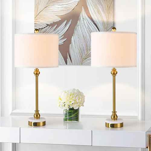 Two matching table lamps with white drum shades and gold-finished stems and bases, displayed on a white console table in a bright interior setting with a floral wall art piece behind them.