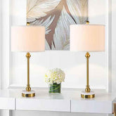 Two matching table lamps with white drum shades and gold-finished stems and bases, displayed on a white console table in a bright interior setting with a floral wall art piece behind them.