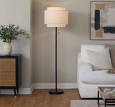 A modern two-tier drum floor lamp with a black metal frame and cream fabric shades, styled in a contemporary living room next to a white sofa and a sideboard.