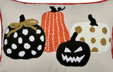A close-up of a decorative textile or pillow featuring three stylized pumpkins: a black pumpkin with white polka dots and a gold bow, an orange pumpkin with white vertical stripes, and a white pumpkin with gold polka dots and a black stem, all on a beige background with red border trim.